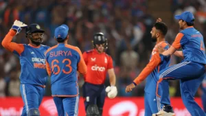 Indian cricket players are celebrating a wicket during a match against England. The players are wearing blue and orange jerseys with "DREAM INDIA" branding. The English batsman in the background, wearing a red jersey, appears disappointed. The scene is set in a stadium with a lively crowd in the background.